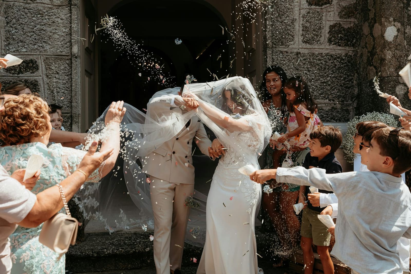 A newly married couple showered with confetti as they leave a stone chapel.