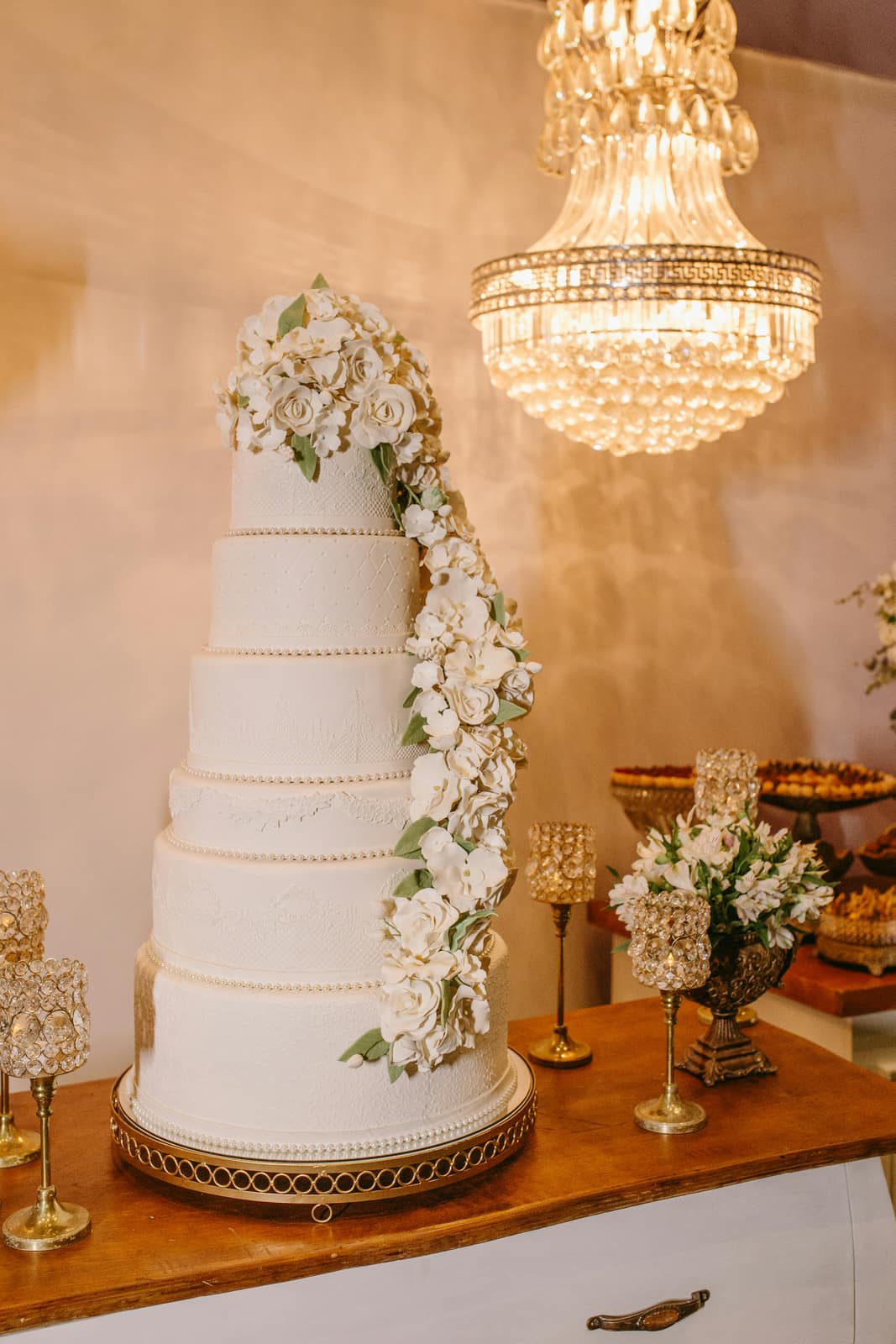 A tiered wedding cake dressed with cascading flowers beneath a chandelier